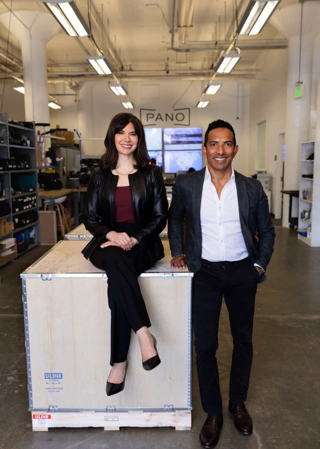 Two people pose in an industrial-style workspace; one sits on a large wooden crate, the other stands beside it. Shelves with equipment and a "PANO" sign are visible in the background, evoking the feel of authentic testimonials in action.